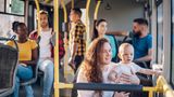 Passengers seated and standing inside a public bus with yellow handrails, including an adult holding a baby in the foreground while other riders sit and stand nearby.