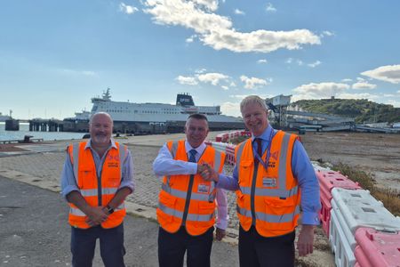 Toby Howe with Peter Osborne and Doug Bannister wearing orange high-visibility vests with two shaking hands. A DFDS ferry, port infrastructure and the white cliffs of Dover are visible in the background.