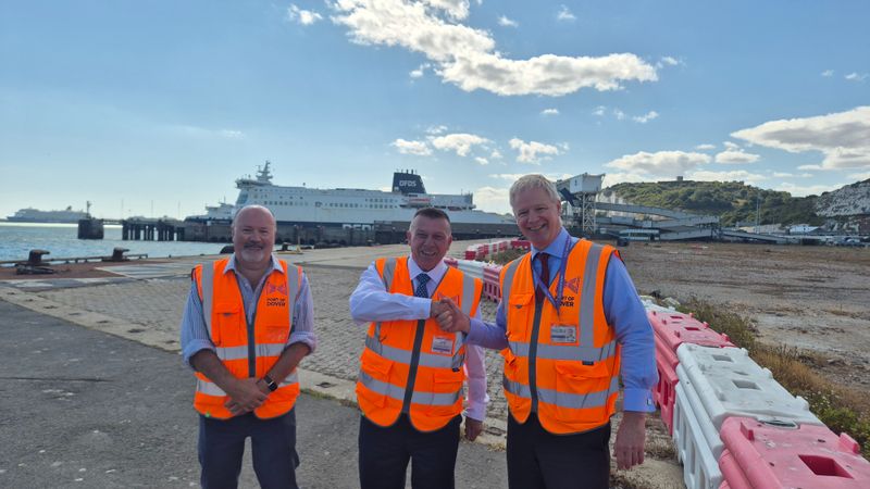 Toby Howe with Peter Osborne and Doug Bannister wearing orange high-visibility vests with two shaking hands. A DFDS ferry, port infrastructure and the white cliffs of Dover are visible in the background.