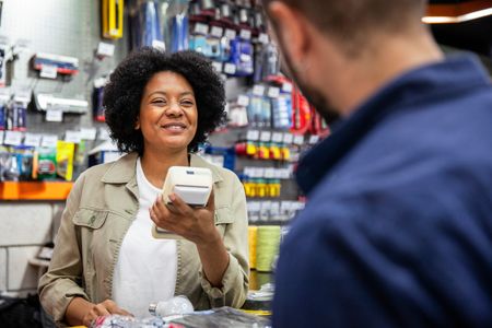 A member of staff at a DIY store checkout holds a card reader while talking to a customer in blue overalls. There are shelves of tools and DIY accessories in the background.