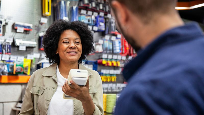 A member of staff at a DIY store checkout holds a card reader while talking to a customer in blue overalls. There are shelves of tools and DIY accessories in the background.