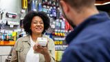 A member of staff at a DIY store checkout holds a card reader while talking to a customer in blue overalls. There are shelves of tools and DIY accessories in the background.