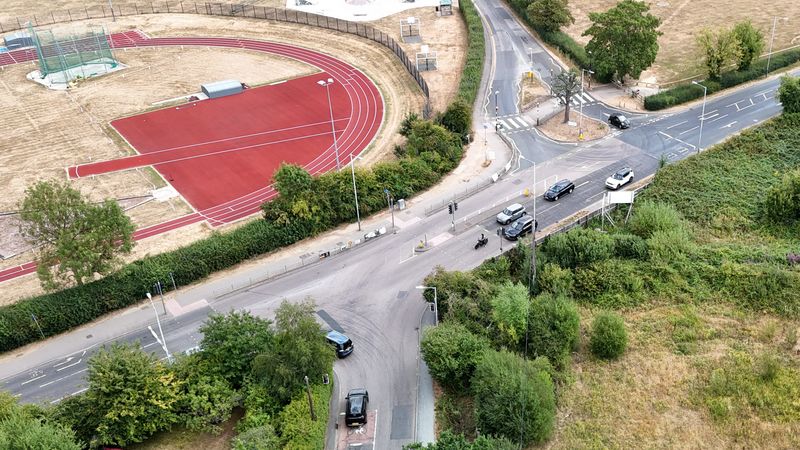 Aerial view of the Badsell Road junction in Paddock Wood, showing the signal-controlled intersection, pedestrian crossing points and several vehicles using the junction. An athletics track is visible to the left, with open land and vegetation surrounding the roads.