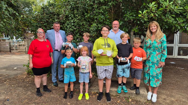 Children and adults from Stones in the Community standing outdoors with handmade model gritters created during a summer club activity. Trees and school buildings can be seen in the background.