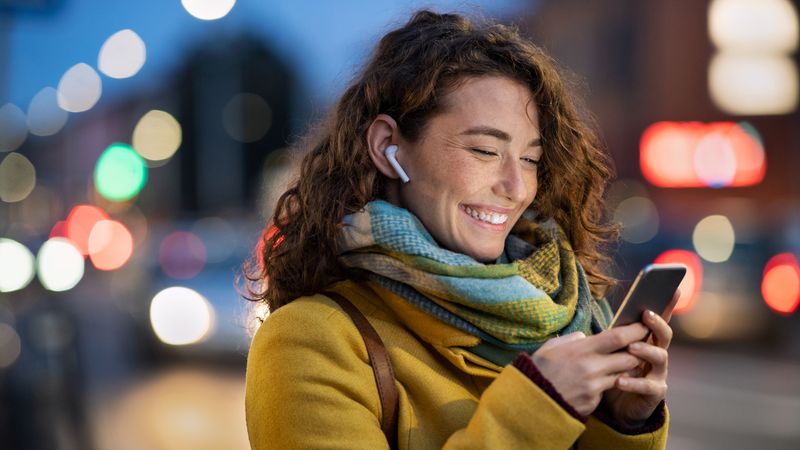 Person wearing a mustard-yellow coat and multicoloured scarf stands on a city street at dusk, looking at a smartphone while wearing wireless earbuds. Blurred vehicle lights and illuminated buildings create a soft-focus urban background.