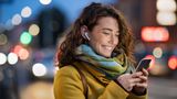 Person wearing a mustard-yellow coat and multicoloured scarf stands on a city street at dusk, looking at a smartphone while wearing wireless earbuds. Blurred vehicle lights and illuminated buildings create a soft-focus urban background.