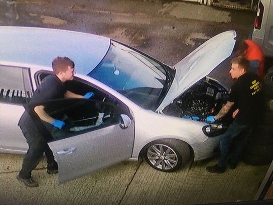 Three individuals are seen working on a silver car. The bonnet is up and the driver's door is open. It is a CCTV still image 