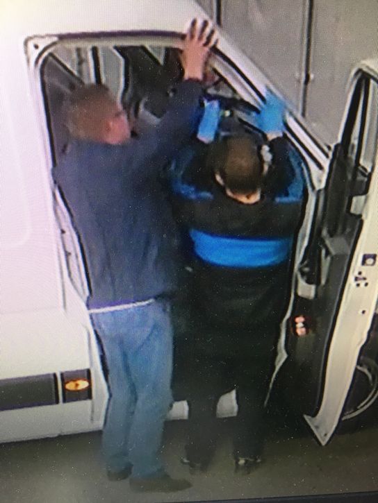 Two individuals work on the dashboard of a white van, back to the camera through the driver's door 