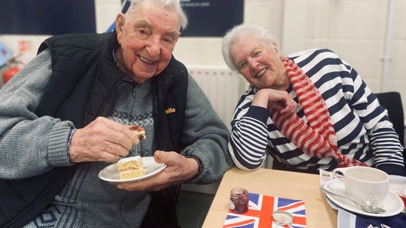 Fred and Joyce, an older couple, sitting at a table and enjoying cakes laid on to mark the 80th anniversary of Victory in Europe. They are smiling