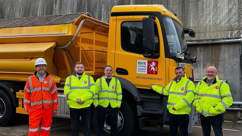 A group of people wearing hi-vis smile towards the camera infront of a big yellow gritter.