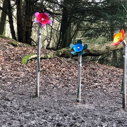 Three colourful metal flower sculptures with bright petals standing in a muddy forest area.