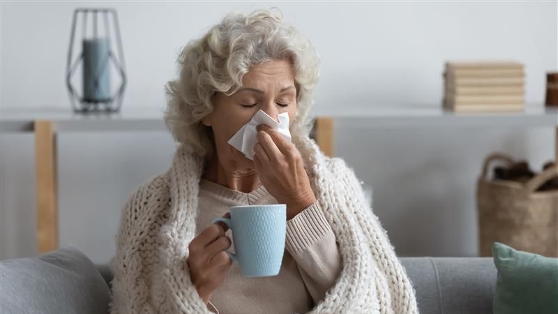 Older woman wrapped in blanket, sat on sofa, holding a hot drink and blowing nose. 