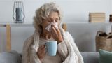 Older woman wrapped in blanket, sat on sofa, holding a hot drink and blowing nose. 