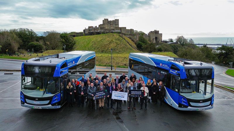 A drone image of a group of people standing looking up. They are standing next to two blue buses and in the background is a castle.