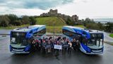 A drone image of a group of people standing looking up. They are standing next to two blue buses and in the background is a castle.