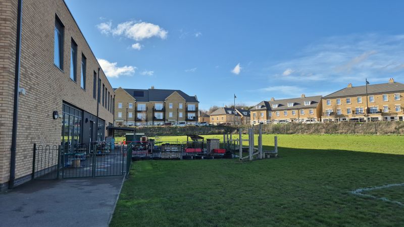Ebbsfleet Green Primary School's main building with a fenced off outdoor play area and equipment sits beside a large grassy field. In the background, rows of modern brick houses stand under a blue sky.