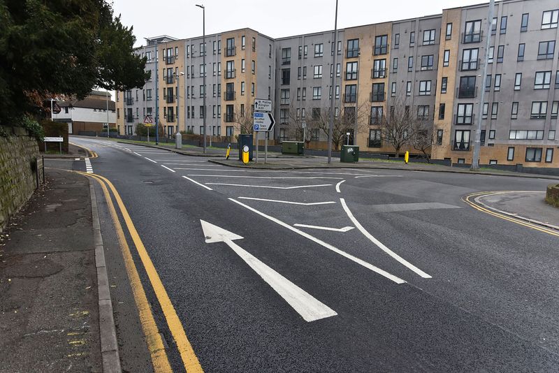An urban junction with fresh white road markings, including an arrow directing traffic straight ahead. The road is bordered by double yellow lines and tall modern apartment buildings stand in the background.