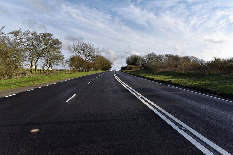 A wide, freshly resurfaced road with solid double white centre lines running uphill through open countryside. Trees line both sides of the road and the sky is bright with scattered clouds