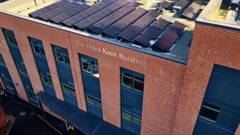 An exterior view of a modern brick building with large dark-framed windows and rows of solar panels covering the roof. Text on the wall reads &lsquo;The Helen Keen Building.&rsquo; The image is taken from an elevated angle.
