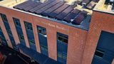 An exterior view of a modern brick building with large dark-framed windows and rows of solar panels covering the roof. Text on the wall reads &lsquo;The Helen Keen Building.&rsquo; The image is taken from an elevated angle.