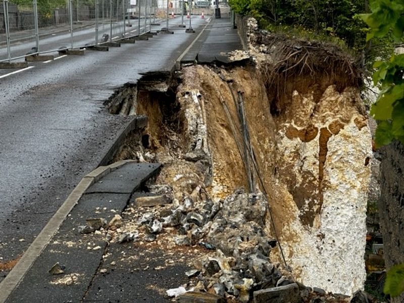 Severe ground collapse beside a road, showing a large exposed pit with broken pavement, soil layers, debris, and visible pipes next to a fenced roadway.