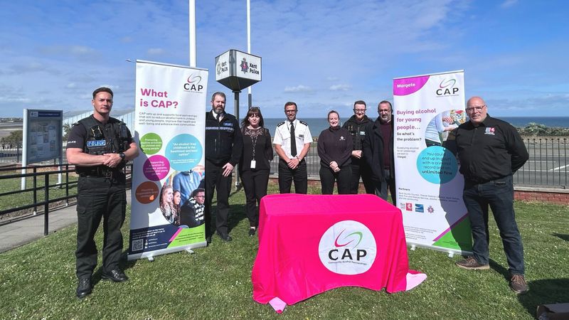 Uniformed officers and community representatives standing on grass near a seafront beside Community Alcohol Partnerships (CAP) banners. In front of the group, in the centre of the photo, is a table covered in a CAP-branded bright pink cloth  