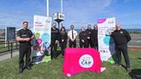 Uniformed officers and community representatives standing on grass near a seafront beside Community Alcohol Partnerships (CAP) banners. In front of the group, in the centre of the photo, is a table covered in a CAP-branded bright pink cloth  