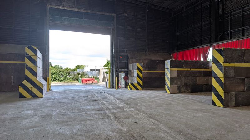 Interior of a waste transfer station showing a newly laid concrete floor, large open vehicle entrance, and reinforced waste bays with black and yellow hazard markings along the walls.