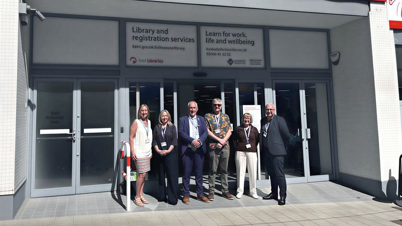 Six people stand outside the entrance of Folkestone Library and Community Learning Centre on a sunny day. Above the glass doors is a sign with the library&rsquo;s name and Kent County Council and Community Learning and Skills logos, while printed panels on the doors advertise library services and adult learning opportunities.