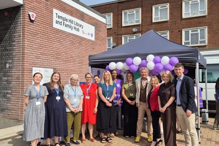 A large group of people pose outside a brick building with a sign reading &ldquo;Temple Hill Library and Family Hub.&rdquo; The group stands in front of a small canopy decorated with purple and white balloons, with nearby buildings and a parked vehicle visible in the background.