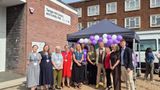 A large group of people pose outside a brick building with a sign reading &ldquo;Temple Hill Library and Family Hub.&rdquo; The group stands in front of a small canopy decorated with purple and white balloons, with nearby buildings and a parked vehicle visible in the background.