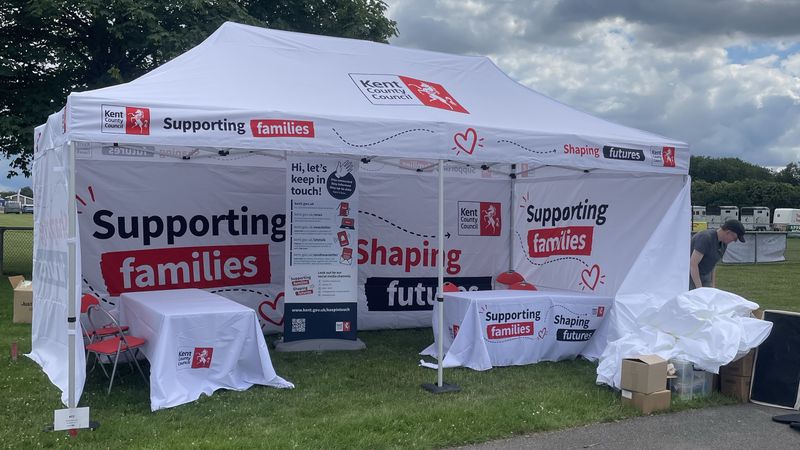 Kent County Council-branded white gazebo at an outdoor event, featuring large &ldquo;Supporting families, Shaping futures&rdquo; graphics on the canopy and walls. The stand contains tables, chairs and an information banner inviting residents to stay in touch. The gazebo is set on grass with trees and cloudy skies in the background, while event equipment and a person setting up are visible to one side.