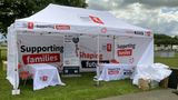 Kent County Council-branded white gazebo at an outdoor event, featuring large &ldquo;Supporting families, Shaping futures&rdquo; graphics on the canopy and walls. The stand contains tables, chairs and an information banner inviting residents to stay in touch. The gazebo is set on grass with trees and cloudy skies in the background, while event equipment and a person setting up are visible to one side.