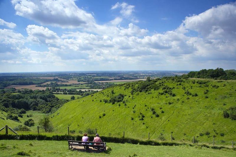 Two people sitting on a bench overlooking the rolling green slopes of the Kent Downs and surrounding farmland under a blue sky with scattered clouds.