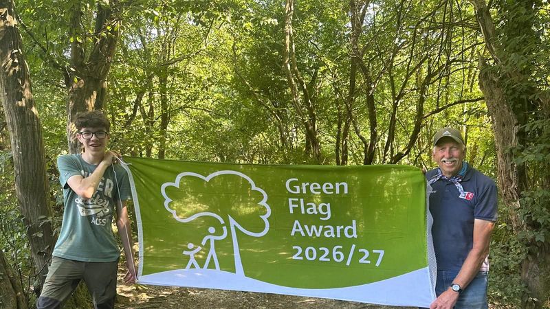 Two men stand in a country park holding up a green flag which says: 'green flag award 26/27