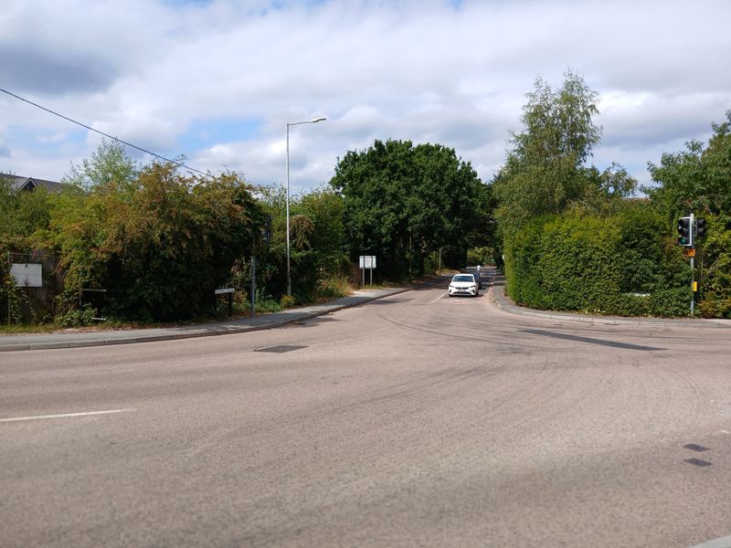 Ground-level view of the Badsell Road junction in Paddock Wood, showing a signal-controlled side road joining a wider main road. Trees and hedges line both sides of the junction, with a car approaching from Badsell Road and traffic lights visible on the corner