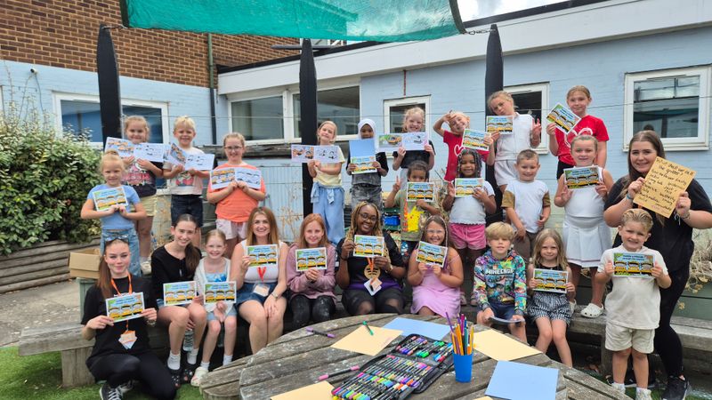 Group of children and activity leaders gathered in an outdoor play area, holding activity books for Kent County Council's gritter naming competition. Art materials are displayed on a table in the foreground.