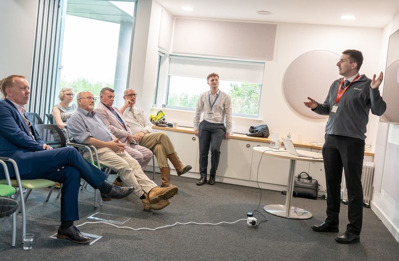 Presentation taking place in a meeting room, with a speaker standing beside a table and laptop while a small audience listens.