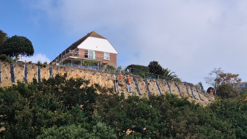 A house sits at the top of a steep cliff above dense vegetation, while workers in high-visibility orange safety clothing carry out stabilisation work on the exposed cliff face. Protective mesh and rock anchors are visible along the slope beneath the property.