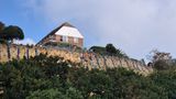 A house sits at the top of a steep cliff above dense vegetation, while workers in high-visibility orange safety clothing carry out stabilisation work on the exposed cliff face. Protective mesh and rock anchors are visible along the slope beneath the property.