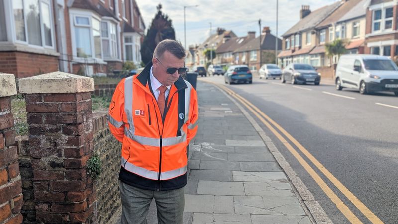 A man wearing an orange hi-viz walks along inspecting  a road.