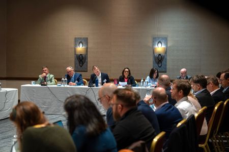 Panel members sit at the front of a conference room during a water partnership meeting, while attendees listen and take part in discussion from tables arranged around the room.