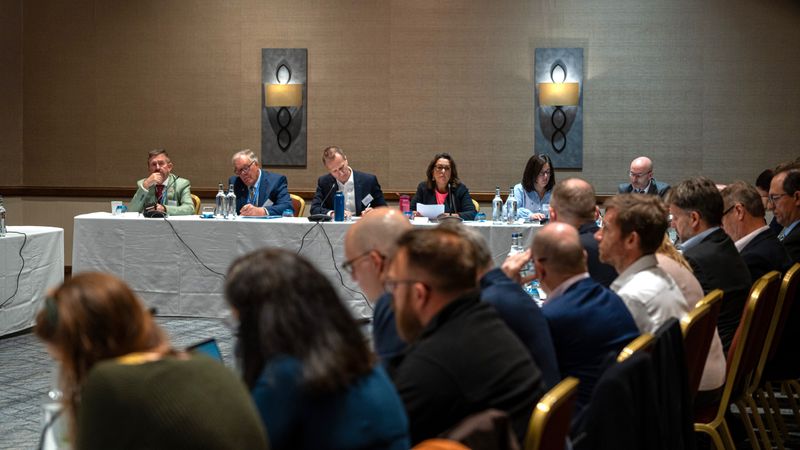 Panel members sit at the front of a conference room during a water partnership meeting, while attendees listen and take part in discussion from tables arranged around the room.