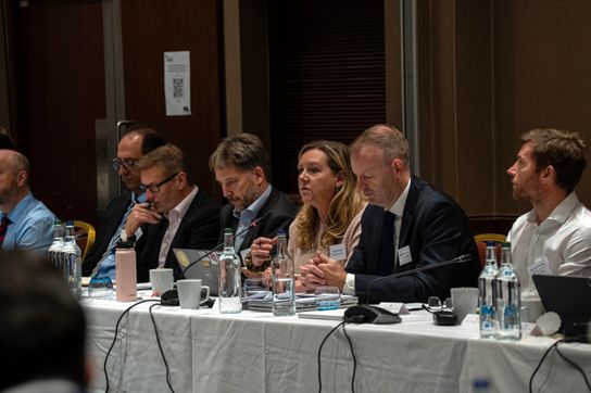 Participants sit at a conference table during a water sector discussion, with one attendee speaking into a microphone while others listen, review documents and make notes. Water bottles, laptops and meeting papers are arranged along the table in a conference room setting.