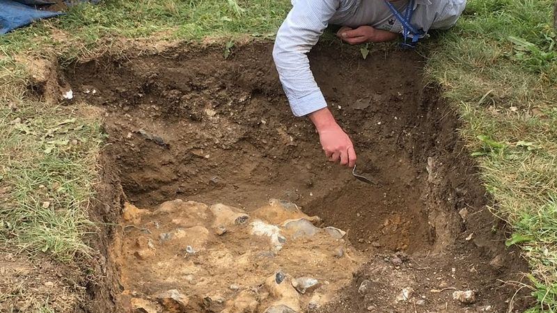 Image of archaeologist Andrew Mayfield laying on the floor, where he has dug up grass to show a large hole, where an excavation site is