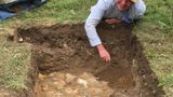 Image of archaeologist Andrew Mayfield laying on the floor, where he has dug up grass to show a large hole, where an excavation site is