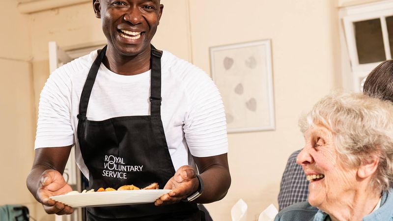 A person in a black Royal Voluntary Service apron serving up a plate of food to a smiling diner