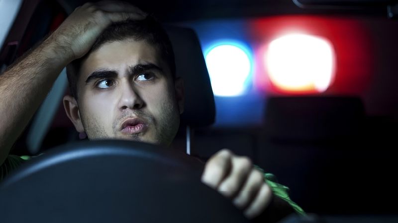A man in a car with his hand on his head looks in his rear-view mirror to see emergency service lights