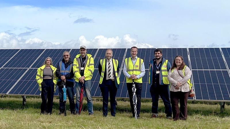 Photograph shows a group of people in a sunny field smiling into the camera. They are in front of solar panels. 