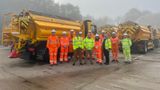 A groupp of people in hi-vis stand in front of yellow gritters